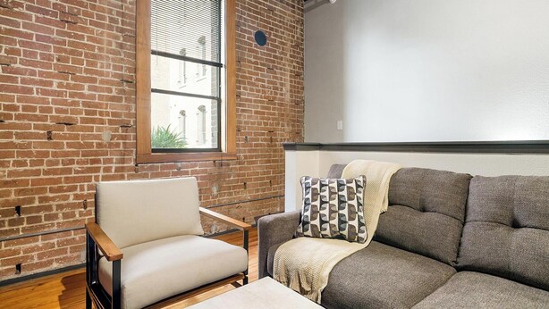 A room with a brick wall, a window, a sofa, a chair, and a white wall. - Landing | Spectacular 1BD, Gym, Courtyard (Houston)