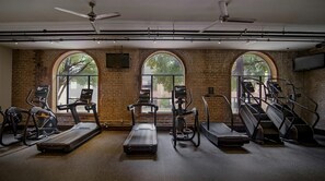 Ein Fitnessstudio mit Laufbändern, Crosstrainern und Gewichtecken, ausgestattet mit großen Bogenfenstern und freiliegenden Backsteinwänden.