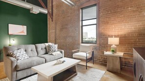 Ein modernes Wohnzimmer mit grüner Wand, Ziegelwand, großem Fenster, grauem Sofa, weißem Sessel, Couchtisch und Beistelltisch mit Lampe.