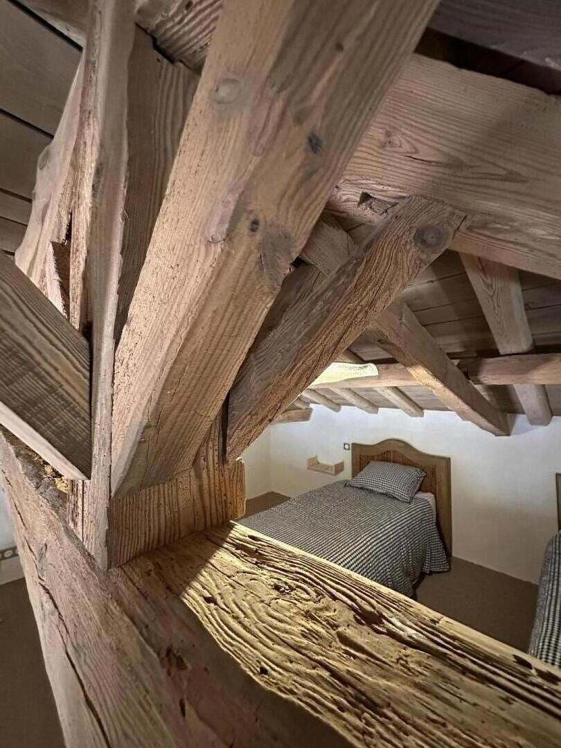 Une structure de plafond en bois avec des poutres apparentes, un lit recouvert d’une couverture à carreaux et une étagère fixée au mur.