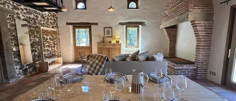 A dining area with a wooden table set for six, a stone wall, a fireplace, and a loft with a staircase.