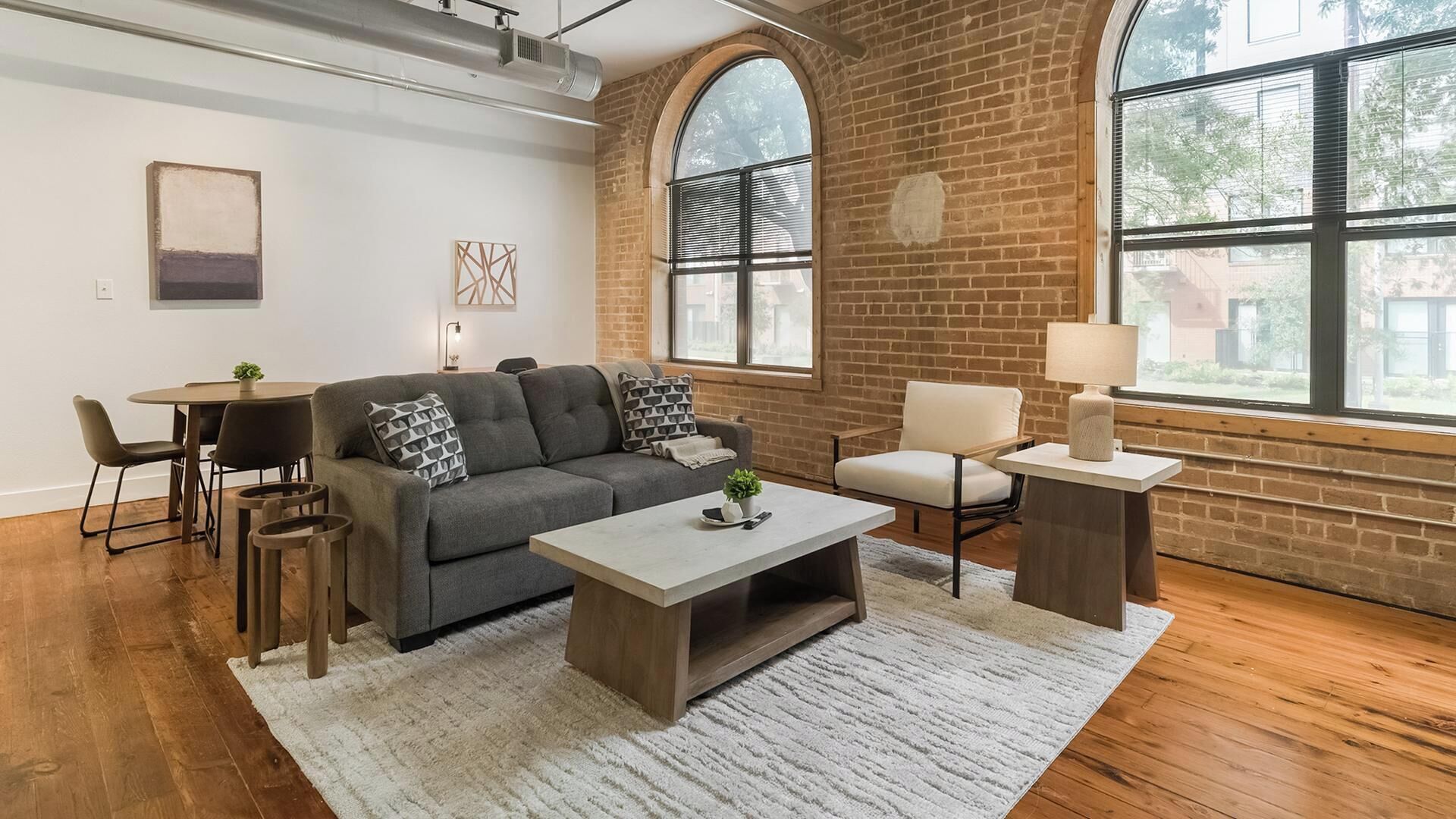 A modern living room with a grey sofa, a coffee table, a dining table with chairs, a white armchair, and a lamp.