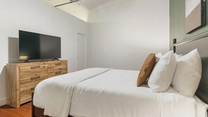 A bedroom with a bed, a wooden dresser, a television, and a ceiling light fixture. - Landing | Stylish 1BD, Gym, Courtyard (Houston)