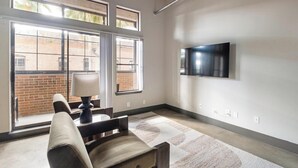 A modern living room with a flat-screen TV mounted on the wall, a sofa, a table with a lamp, and large windows with a view of a brick building. - Landing | Beautiful 1BD, Gym, Courtyard (Houston)