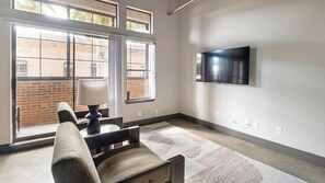 A modern living room with a flat-screen TV mounted on the wall, a sofa, a table with a lamp, and large windows with a view of a brick building. - Landing | Beautiful 1BD, Gym, Courtyard (Houston)