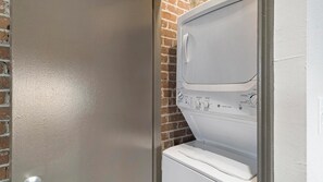 A laundry room with a white washing machine and dryer, a brick wall, and a door. - Landing | Amazing 2BD, Courtyard, Gym (Houston)