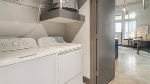 A laundry room with two white washing machines and a dryer, a metal vent above the machines, and a view into a dining area with a table and chairs. - Landing | Incredible 1BD, Gym, Courtyard (Houston)