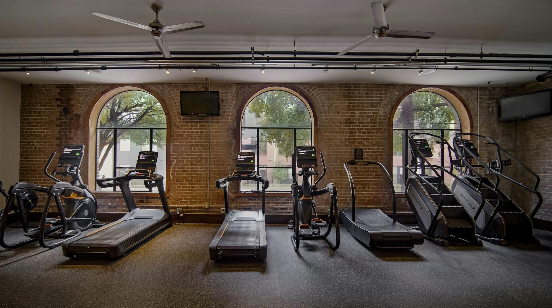 A gym with treadmills, elliptical machines, and weight stations, featuring large arched windows and exposed brick walls.