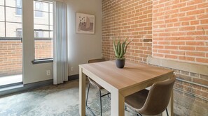 A room with a wooden table, a single chair, a potted plant, a window with blinds, and a brick wall. - Landing | Incredible 1BD, Gym, Courtyard (Houston)