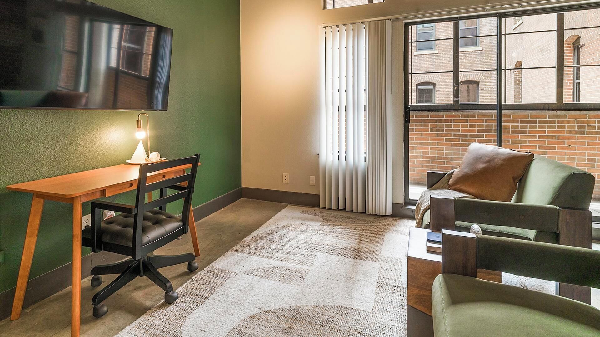 Ein möbliertes Zimmer mit Schreibtisch, Stuhl, Sofa und einem großen Fenster mit Jalousien.