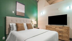 A hotel room with a bed, a television, a nightstand with a lamp, and a wooden dresser. - Landing | Modern 1BD, Courtyard, Gym (Houston)