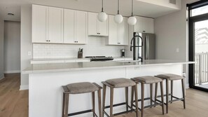 A modern kitchen with white cabinetry, a central island with bar stools, and pendant lighting. - Landing | Beautiful 2BD, Yoga Studio, Gym (Denver)