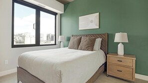 A bedroom with a bed, two bedside tables with lamps, a window with a view of buildings, and a wall-mounted artwork. - Landing | Beautiful 2BD, Yoga Studio, Gym (Denver)