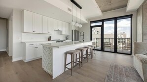 A modern kitchen with white cabinetry, a central island with bar stools, and a large sliding glass door leading to a balcony. - Landing | Stylish 2BD, Gym, Yoga Studio (Denver)