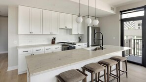 A modern kitchen with white cabinetry, a large island with bar stools, and a view of the outside through a glass door. - Landing | Beautiful 2BD, Yoga Studio, Gym (Denver)