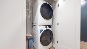 A compact, white laundry room with a stacked washer and dryer, a blue trash bin, and a white door. - Landing | Stylish 2BD, Gym, Yoga Studio (Denver)