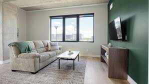 A living room with a sofa, a coffee table, a TV on the wall, and a large window with a view of buildings. - Landing | Stylish 2BD, Gym, Yoga Studio (Denver)