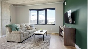 Ein Wohnzimmer mit einer Couch, einem Couchtisch, einem an der Wand befestigten Fernseher und einem großen Fenster mit Blick auf Gebäude.