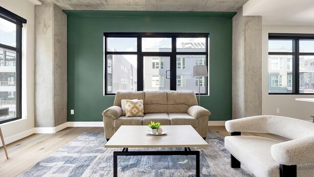 Een moderne woonkamer met een groene accentmuur, een beige bank, een witte salontafel en uitzicht op gebouwen door grote ramen.
