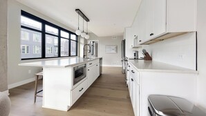 Een moderne keuken met een groot eiland, witte kasten en een uitzicht op gebouwen door grote ramen.