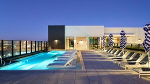 En pool på taket med solstolar och parasoller, en modern byggnad med stora fönster och en utsikt över staden i skymningen.