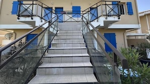 Escadas externas que levam a um edifício com persianas azuis e guarda-corpo de vidro.