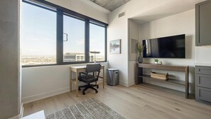 A modern office space with a large window, a desk with a chair, a trash bin, a television, and a shelf with decorative items. - Landing | Stunning 1BD, Clubhouse, Rooftop Lounge (Denver)