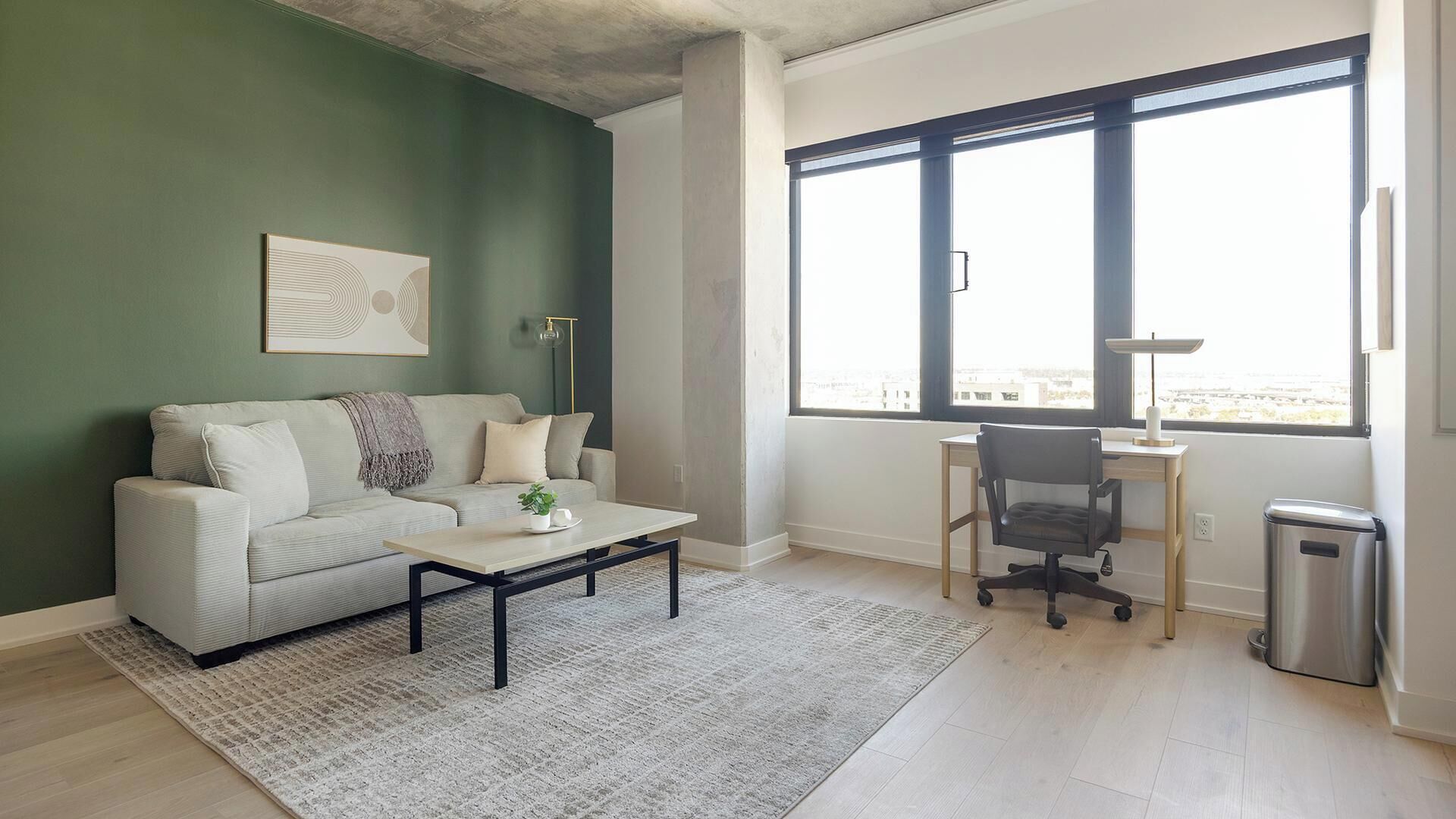 Ein Wohnzimmer mit Sofa, Couchtisch und Schreibtisch mit Stuhl. Ein großes Fenster bietet einen Ausblick nach draußen.