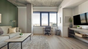 A modern living room with a sofa, coffee table, and a desk with a chair. There's a large window with a view, a TV on the wall, and a trash can. - Landing | Incredible 1BD, Yoga Studio, Clubhouse (Denver)