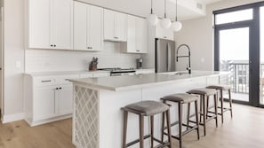 A modern kitchen with white cabinetry, a large island with bar stools, a stainless steel refrigerator, and a view through a glass door. - Landing | Amazing 2BD, Yoga Studio, Gym (Denver)