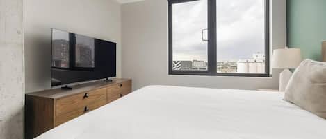 Ein Hotelzimmer mit einem Bett, einem Fernseher auf einer Holzkommode, einem Fenster mit Blick auf Gebäude und einer Lampe auf dem Nachttisch.