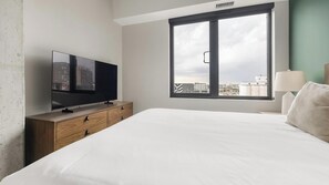 Hotellihuone, jossa on sänky, puinen lipasto, jonka päällä on televisio, ikkuna, josta on näkymä rakennuksiin, ja yöpöydällä oleva lamppu.