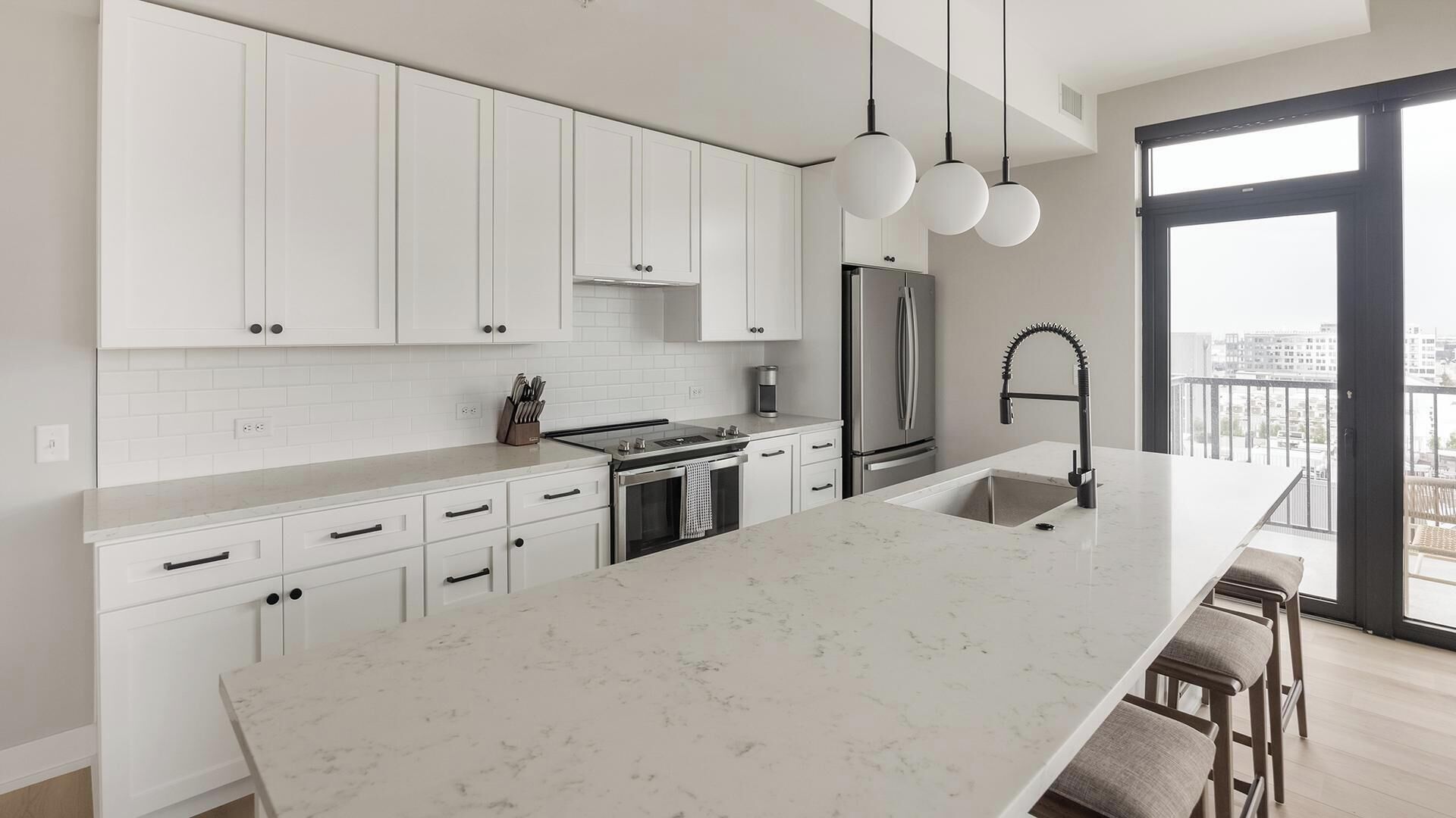 Une cuisine moderne avec des meubles blancs, un grand îlot central, des appareils en acier inoxydable et des luminaires suspendus.