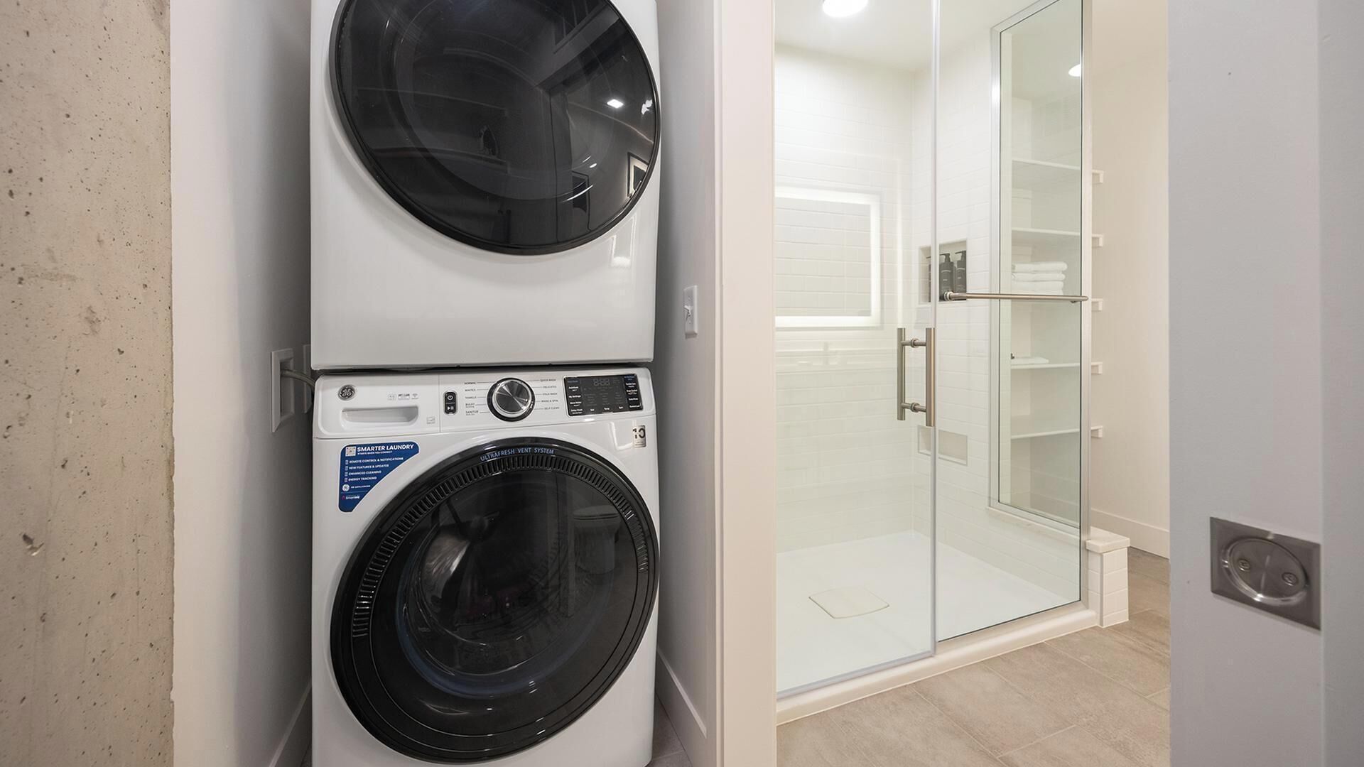 A compact laundry area with a front-loading washer and dryer, a glass shower enclosure, and a tiled floor.