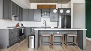 A modern kitchen with grey cabinets, a stainless steel refrigerator, a built-in oven, and a central island with bar stools. - Landing | Chic 1BD, Clubhouse, Yoga Studio (Denver)