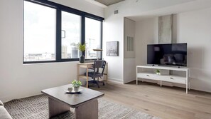 A modern living room with a large window, a flat-screen TV, a desk, and a chair.