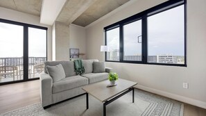 A modern living room with a grey sofa, a coffee table, and a view of the city through large windows. - Landing | Spectacular 2BD, Gym, Clubhouse (Denver)
