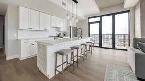 A modern kitchen with white cabinets, a central island with bar stools, and a large sliding glass door leading to a balcony. - Landing | Stunning 2BD, Clubhouse, Yoga Studio (Denver)