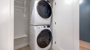 A compact, wall-mounted washer and dryer unit in a small laundry room with a plain wall and a wooden floor. - Landing | Spectacular 2BD, Gym, Clubhouse (Denver)