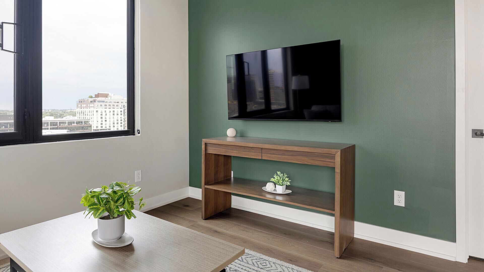 Una habitación con una televisión montada en una pared verde, una mesita de consola de madera con una planta y una ventana con vistas a edificios.