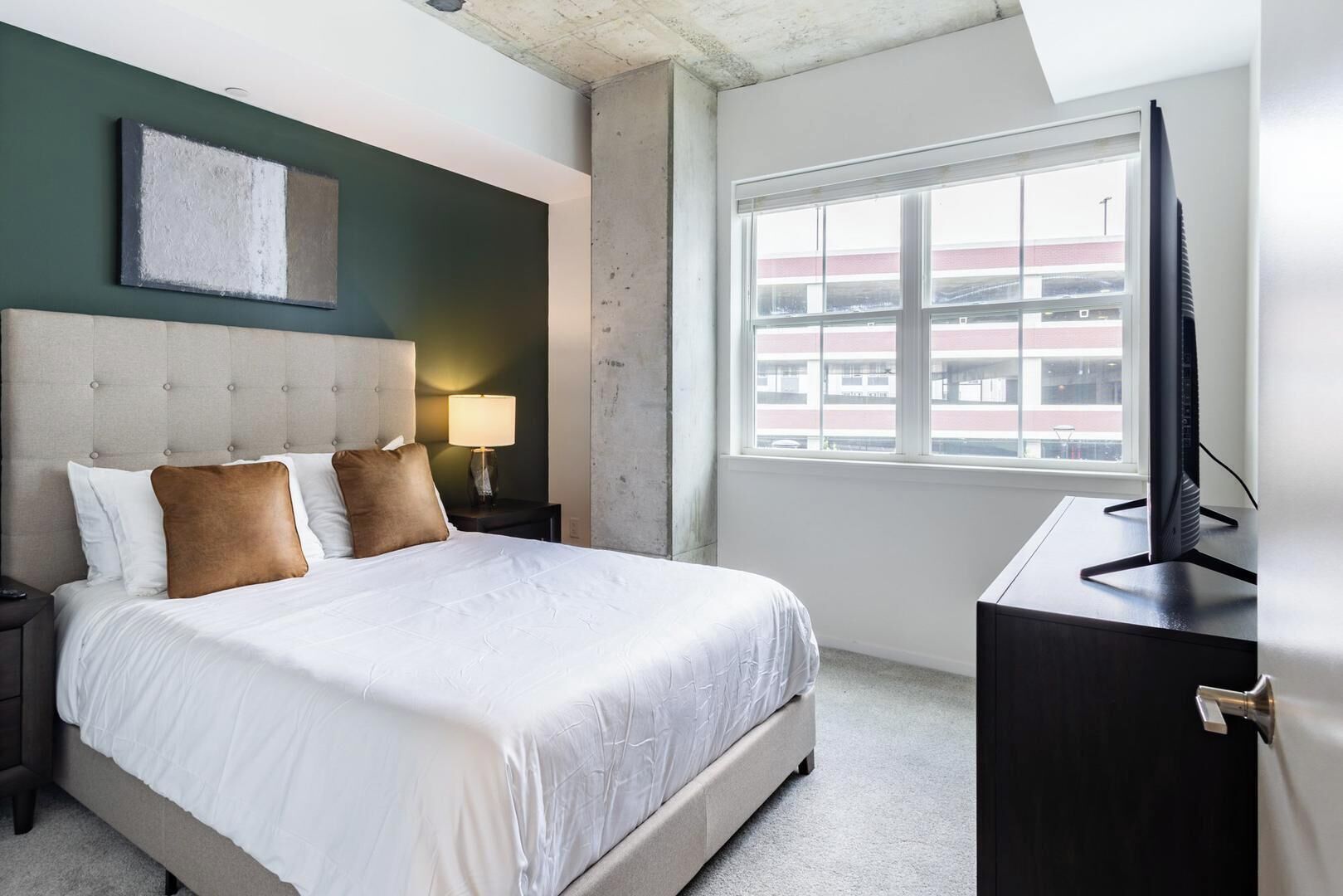 Ein Hotelzimmer mit Bett, Nachttisch, Lampe, Fernseher und einem Fenster mit Blick auf Gebäude.
