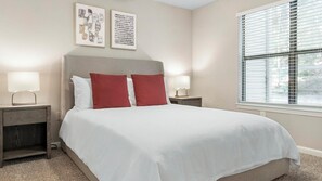 Una cama bien hecha con almohadas blancas y rojas, dos mesitas de noche con lámparas, y una obra de arte en la pared.