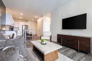 A modern living room with a flat-screen TV, a dining area with a table and chairs, and a kitchen with cabinets and a refrigerator.