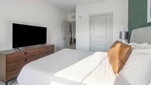 A hotel room with a bed, a TV on a wooden dresser, a door, and a wall-mounted lamp. - Landing | Beautiful 2BD, Gym (St. Augustine)