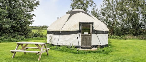 A yurt with a door, a picnic table, and a grassy field with trees in the background.