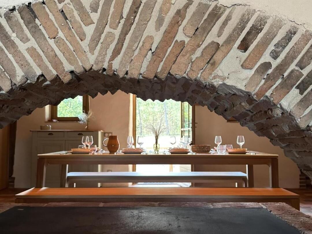 Uma área de refeições com arco de tijolos, mesa de madeira, bancos e uma janela com vista.