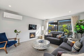 A modern living room with a blue chair, grey sofa, round coffee table, fireplace, TV, and large windows with a view of greenery. - Base Northlake | Peaceful with Mountain Views (Wānaka)