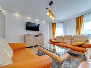 Un salon moderne avec des canapés en cuir orange, une table basse en verre, un téléviseur à écran plat, des meubles en bois et des rideaux jaunes.