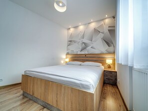Une chambre à coucher moderne avec un lit en bois, des œuvres d’art géométriques au mur, des spots encastrés au plafond et une table de chevet avec une lampe.