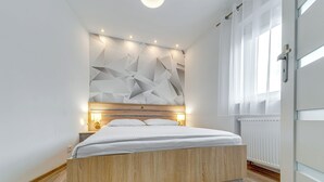 A modern bedroom with a wooden bed, geometric wall design, and a window with curtains. - Apartament nr 32 by Interhome (Gliwice)